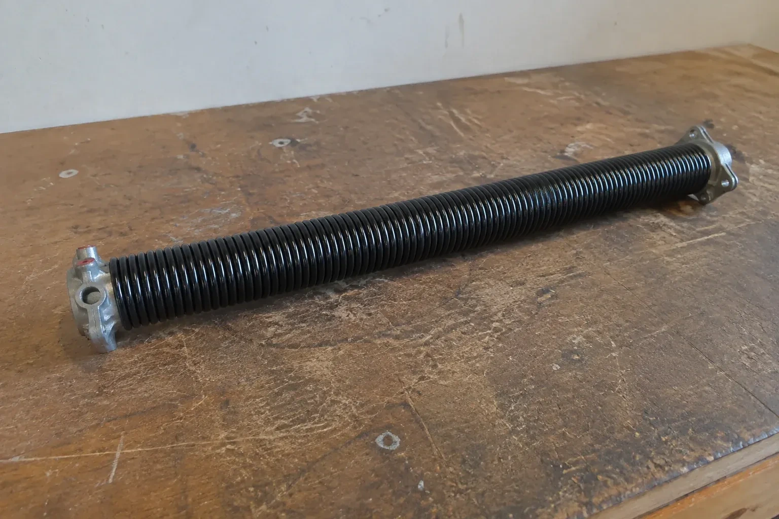 Detached black coil spring with metal end fittings on a workbench. — Torsion Spring Replacement, London