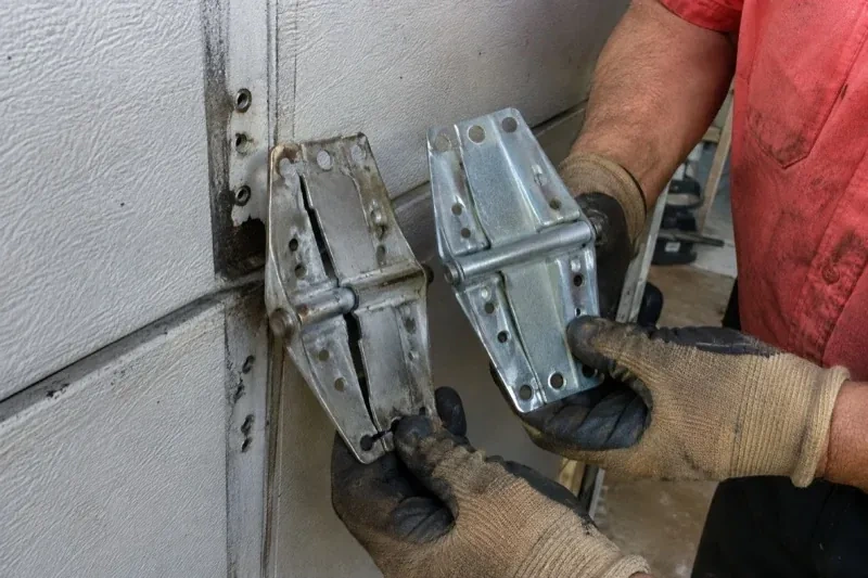 Gloved hands holding two metal garage-door hinges side by side, one discolored with a visible crack and the other bright silver. — Garage Door Hinge Replacement, London