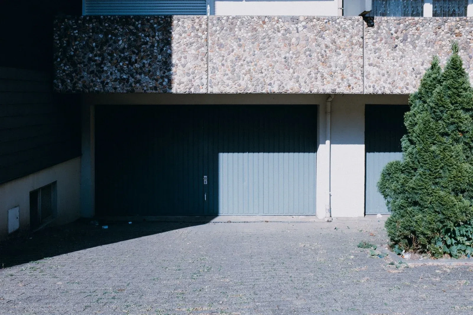 Two gray-blue vertical-plank garage doors under a pebble-textured concrete overhang, half in shadow, beside a green shrub and a paved lot. — White Hills