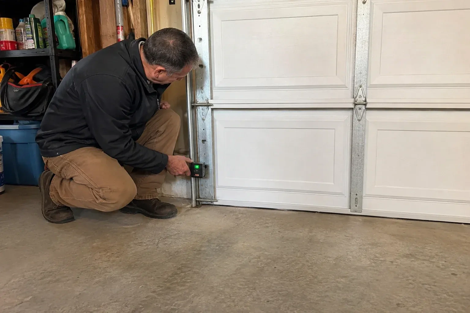 Technician kneeling beside a white paneled garage door, holding a small black box with a green light against the vertical track. — Photo-Eye Sensor Alignment or Replacement, London